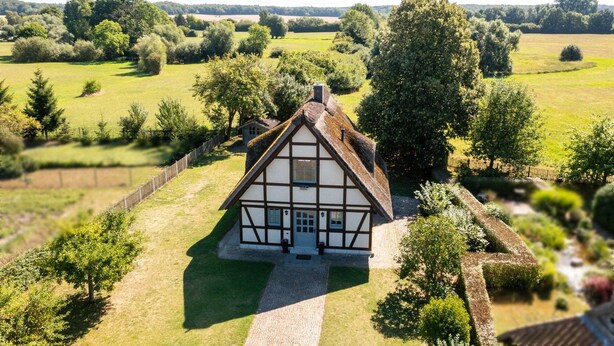 Een klassiek vakwerkhuis met rieten dak, omgeven door een verzorgde tuin en velden.