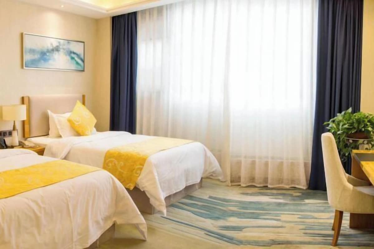 Une chambre d’hôtel avec deux lits, un bureau, une chaise, une plante et un tableau au mur.