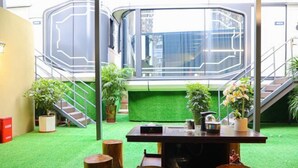 Un área de recepción con piso de césped artificial verde, troncos de madera, una mesa de recepción y una escalera con diseño geométrico.