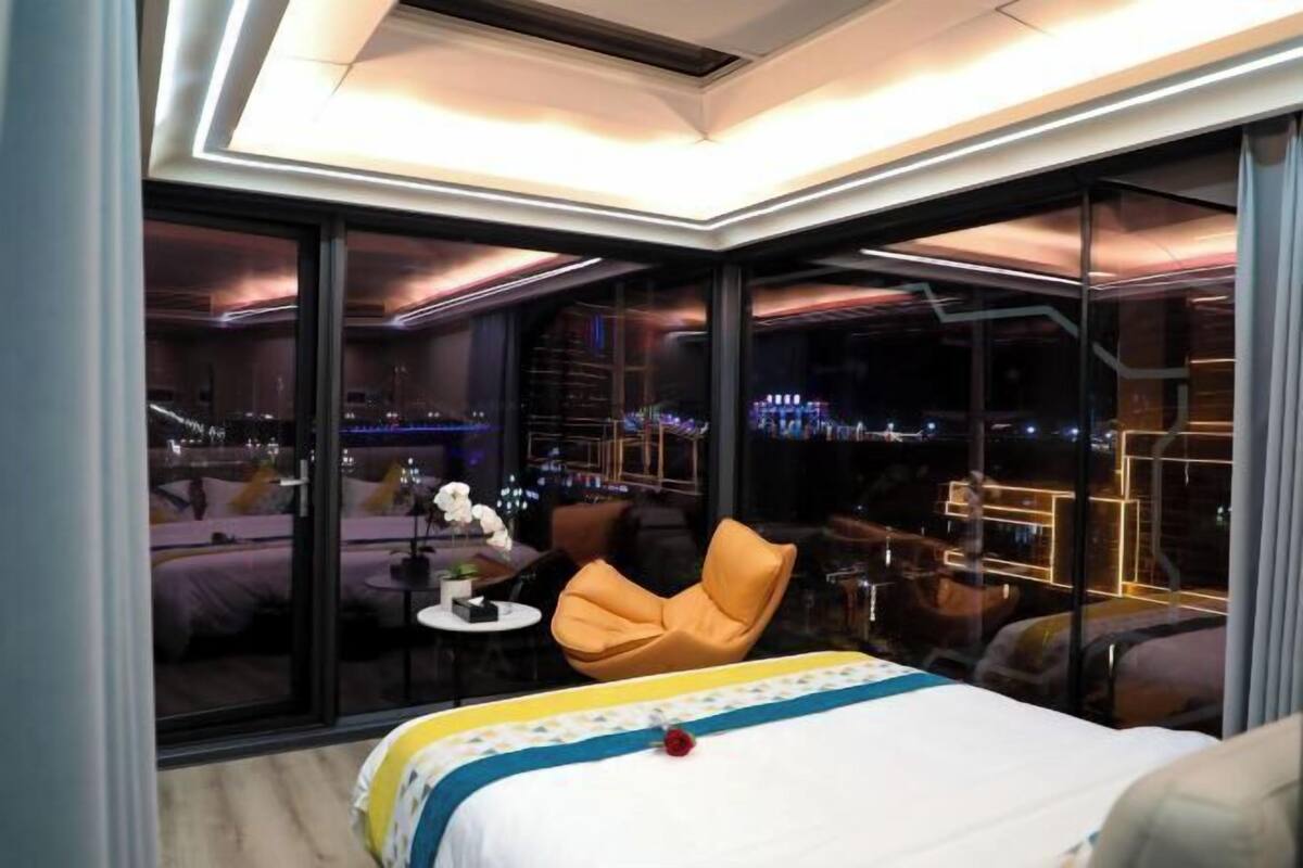 Una habitación de hotel moderna con una cama grande, una mesita redonda pequeña, un sillón cómodo y vistas de la ciudad a través de ventanales del suelo al techo.