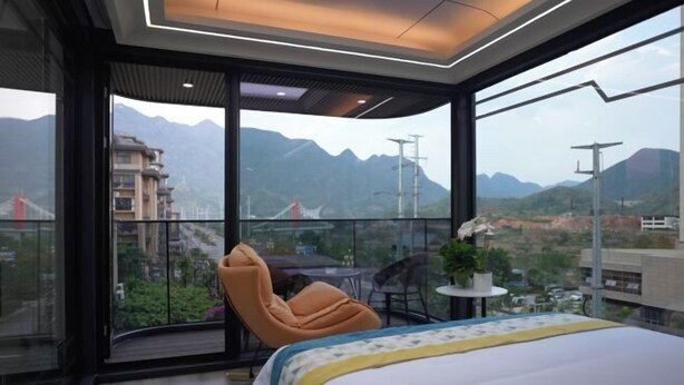 Una habitación de hotel moderna con una cama grande, una silla, una mesita y vistas a montañas a través de amplios ventanales.