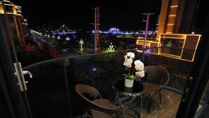 Un balcón con sillas de mimbre y una pequeña mesa, con vistas a un paisaje urbano con edificios iluminados y un puente, de noche.