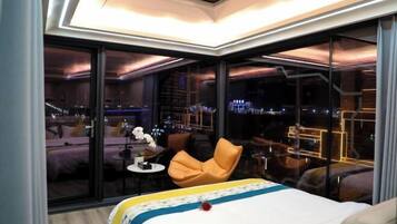 Habitación de hotel moderna con una cama grande, una mesita pequeña, una silla y vistas a la ciudad a través de amplias ventanas.