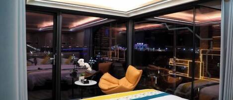 Habitación de hotel moderna con una cama grande, una mesita pequeña, una silla y vistas a la ciudad a través de amplias ventanas.