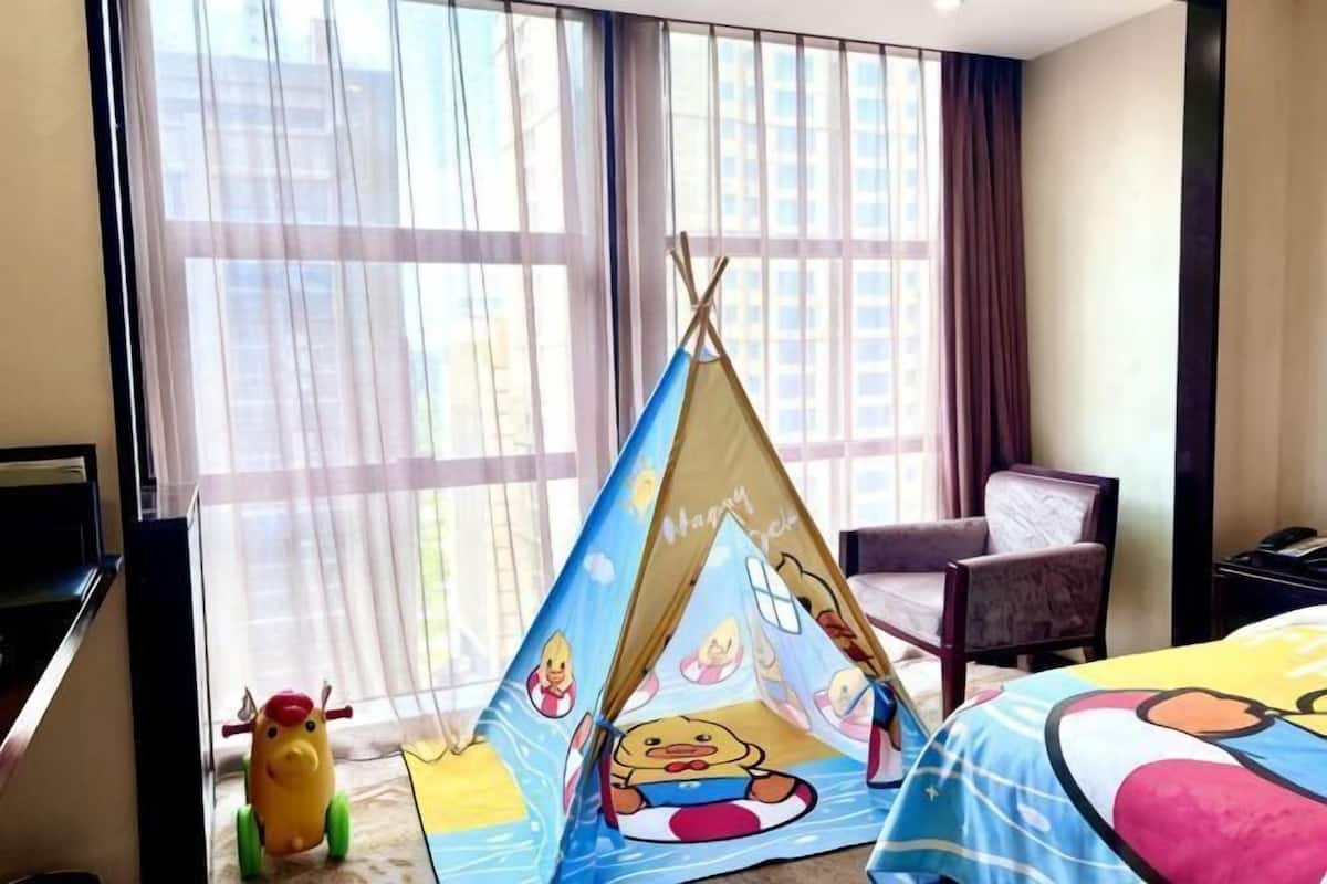 Une chambre d’hôtel avec une grande fenêtre, un lit recouvert d’une courtepointe aux couleurs vives, une chaise et une petite structure en forme de tente décorée de personnages de dessins animés.