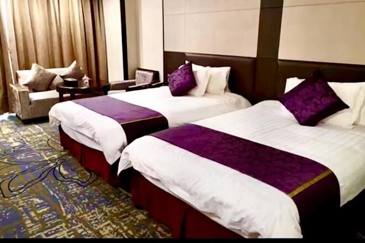 Une chambre d’hôtel avec deux lits, chacun recouvert d’une couvre-lit violette et doté de draps blancs. On y trouve un fauteuil rouge et une petite table avec une lampe. La chambre est agrémentée d’un tapis à motifs et d’une grande fenêtre avec des rideaux.