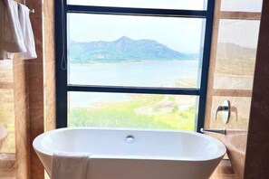 A bathroom with a freestanding bathtub, a window with a view of a lake and mountains, and a towel rack. - Yantai Baohua Mingdu Hotel (Qixia)