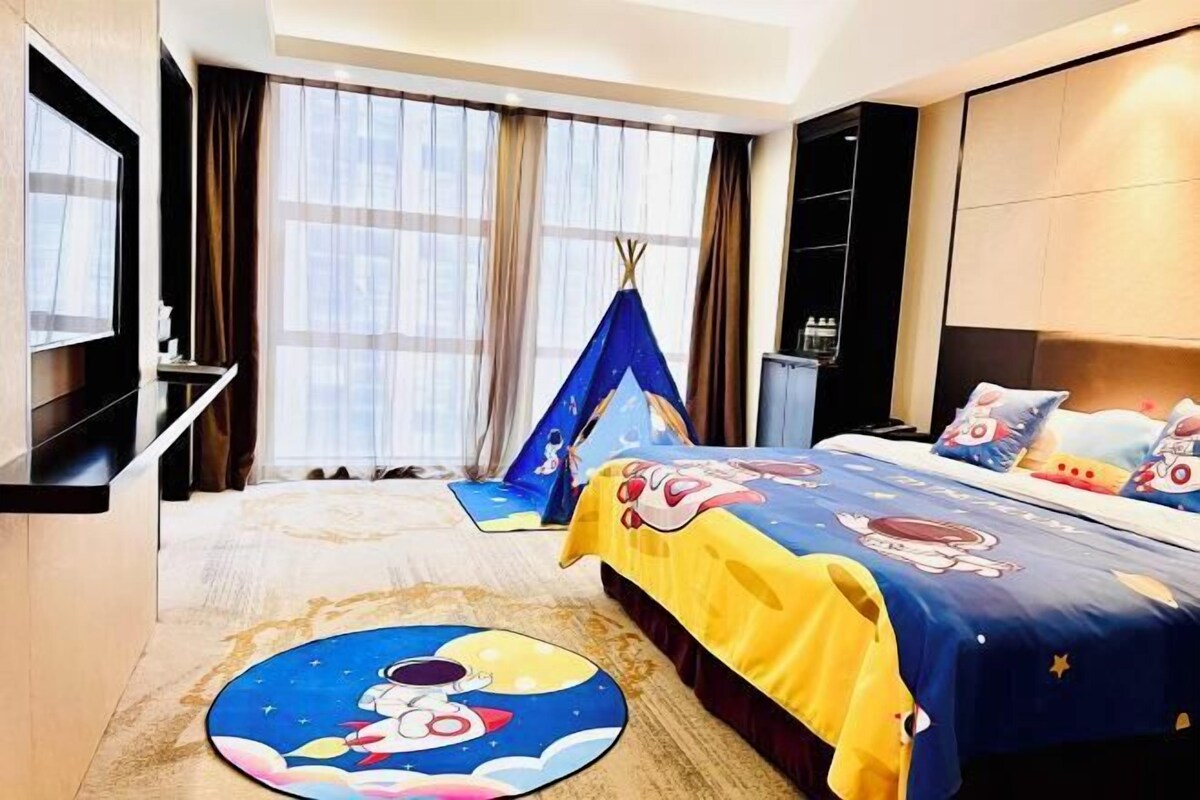 A hotel room with a bed, a TV, a window with curtains, a small table, and a wall-mounted shelf.
