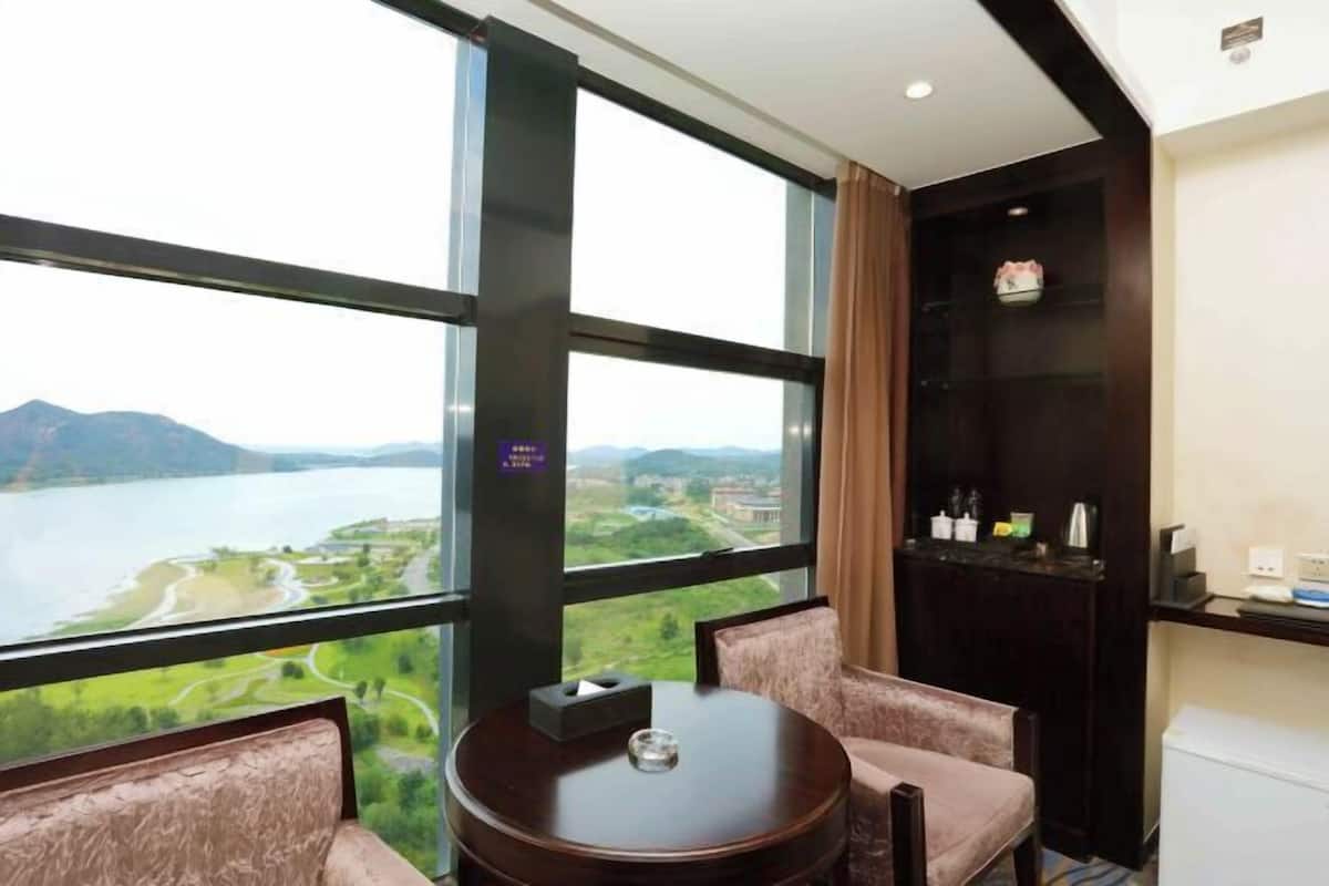 Une chambre d’hôtel dotée d’une grande fenêtre donnant sur un lac et des montagnes, d’une table ronde avec des chaises et d’un mini-bar.