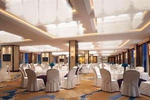 Een feestzaal met ronde tafels en stoelen, een vloerbedekking en een hoog plafond met verlichtingsarmaturen.