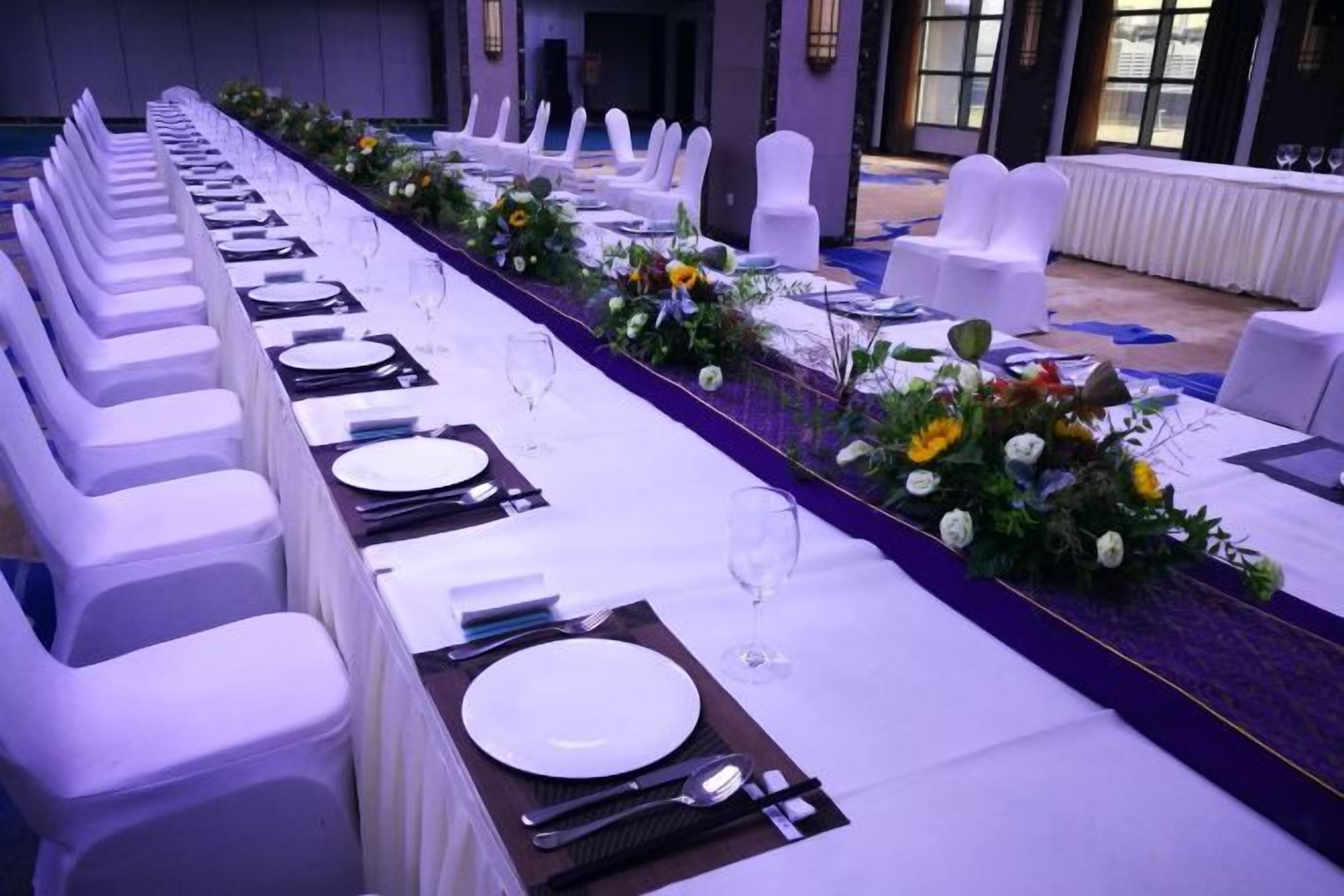 A long banquet table with white chairs, set with plates, cutlery, and glasses, and decorated with floral arrangements.