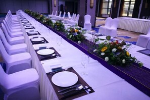 Een lange feesttafel met witte stoelen, gedekt met borden, bestek en glazen, en versierd met bloemarrangementen.