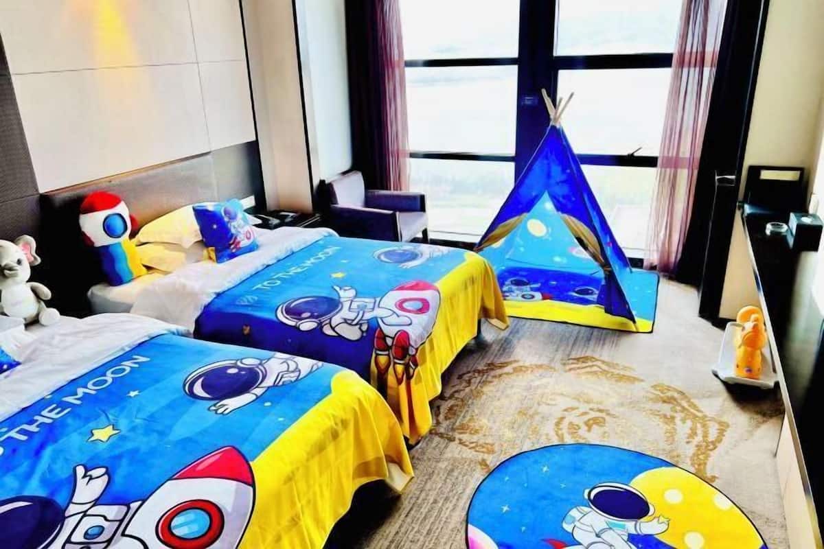 Une chambre d’hôtel avec deux lits, chacun recouvert d’une courtepointe sur le thème de l’espace, une petite table avec un ours en peluche, une fenêtre avec des rideaux et une vue sur le ciel.