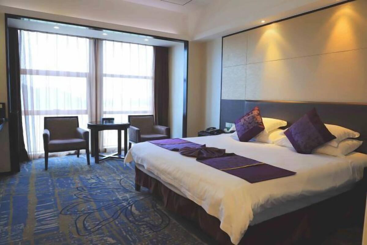 Une chambre d’hôtel avec un grand lit, un bureau et une chaise. Il y a une fenêtre avec des rideaux, une table de chevet et une lampe.