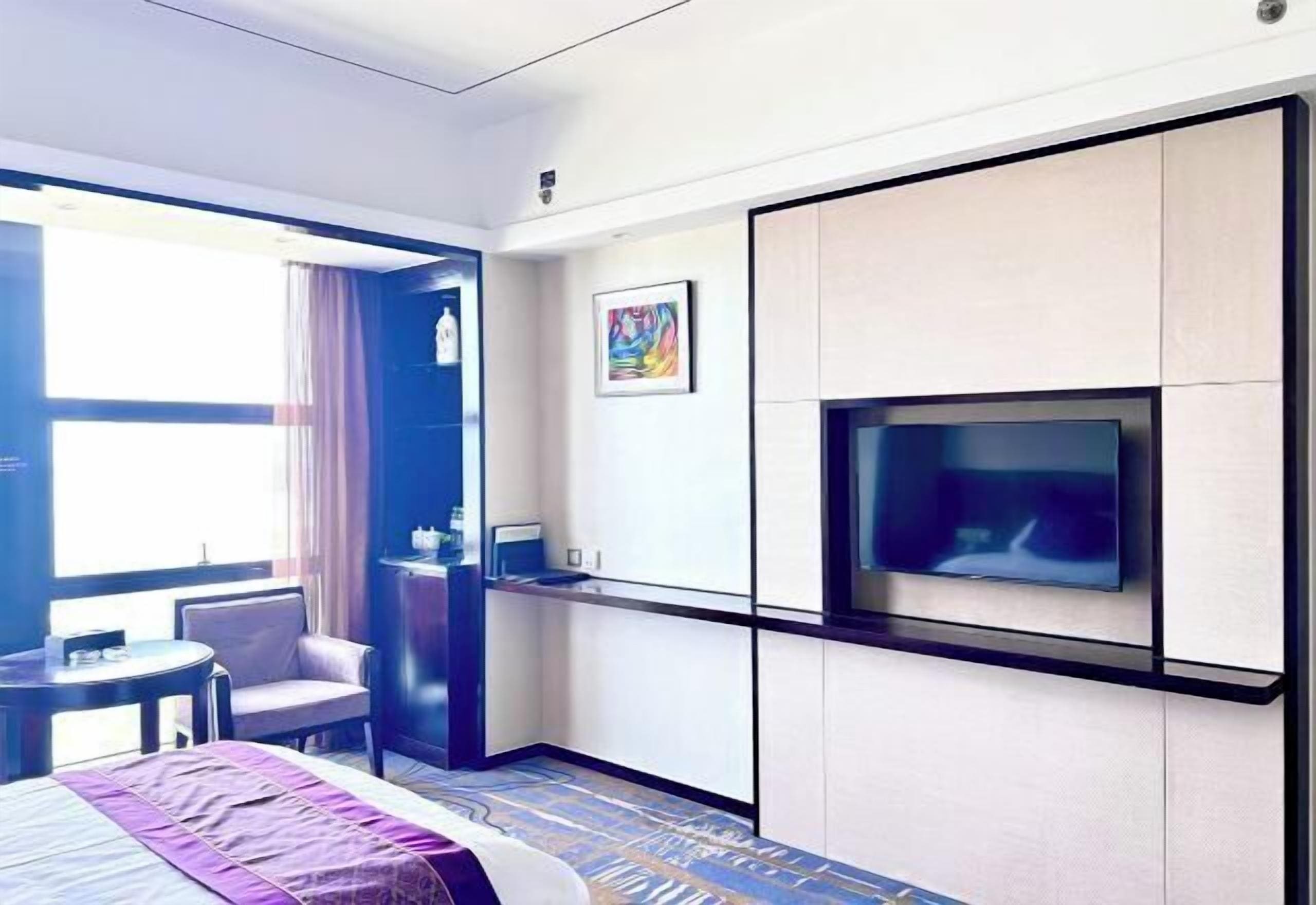 A hotel room with a bed, a TV mounted on the wall, a desk with a chair, and a window with curtains.