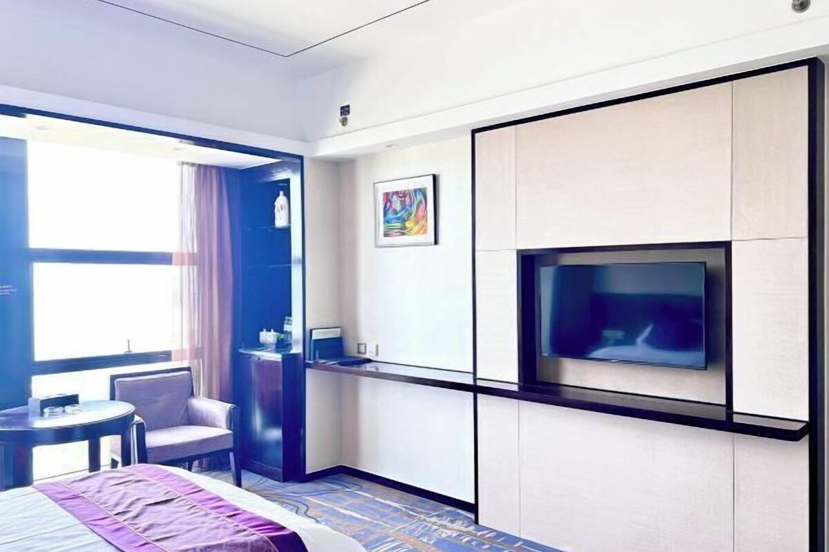 A hotel room with a bed, a TV mounted on the wall, a desk with a chair, and a window with curtains.