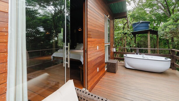 Eine Holzterrasse mit einer Glastür, die zu einem Schlafzimmer führt, einer weißen Badewanne und einem Geländer aus Holz.