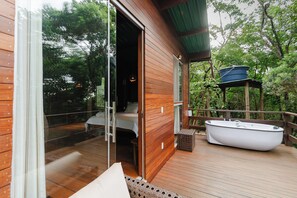 Une terrasse en bois avec une porte vitrée donnant sur une chambre, une baignoire blanche et une rambarde en bois.