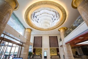 Un vestíbulo de hotel con una gran araña circular, altas columnas y un mostrador de recepción.