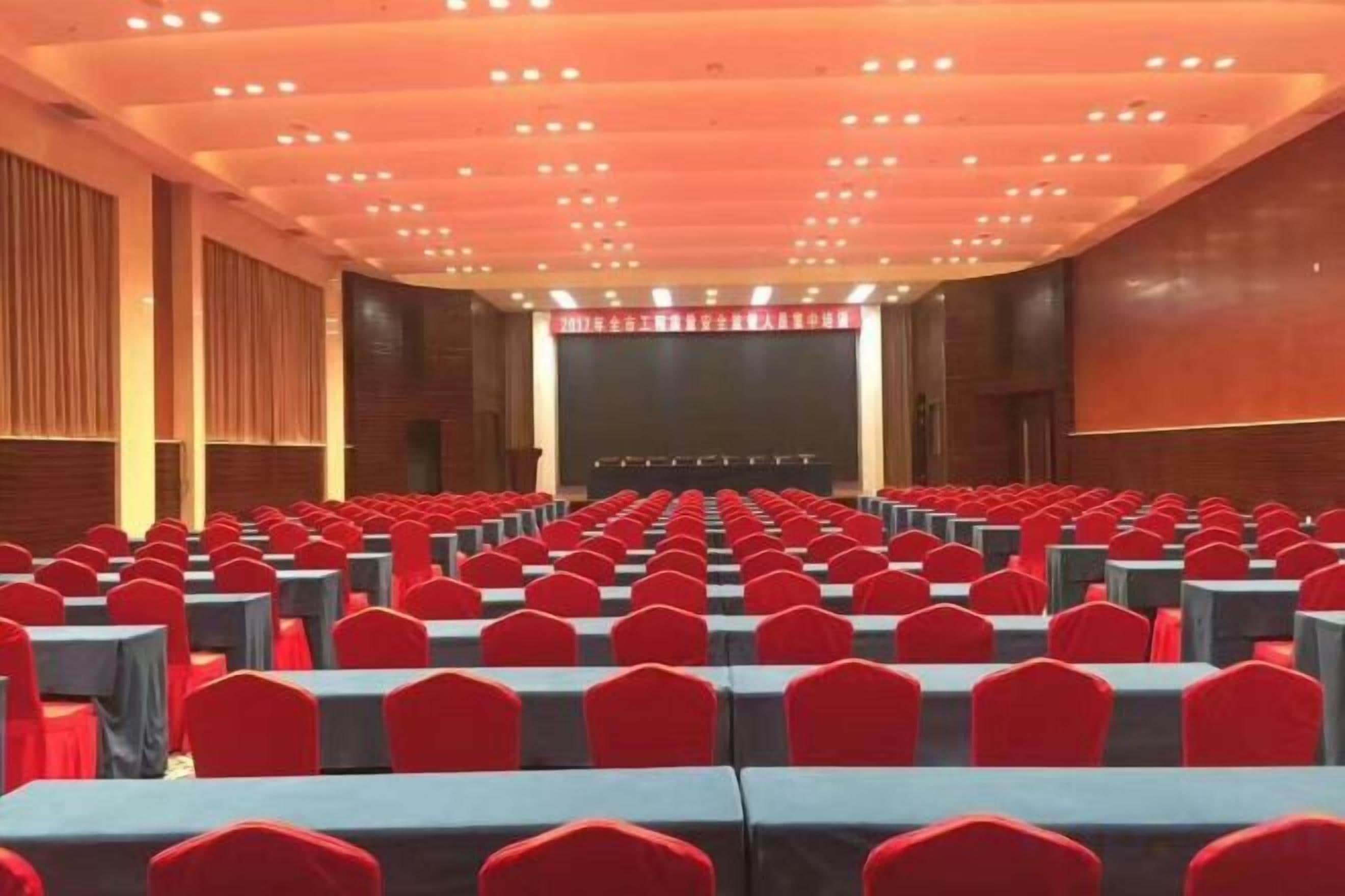 Un salón de conferencias con filas de mesas y sillas, un escenario al frente y una pantalla grande.