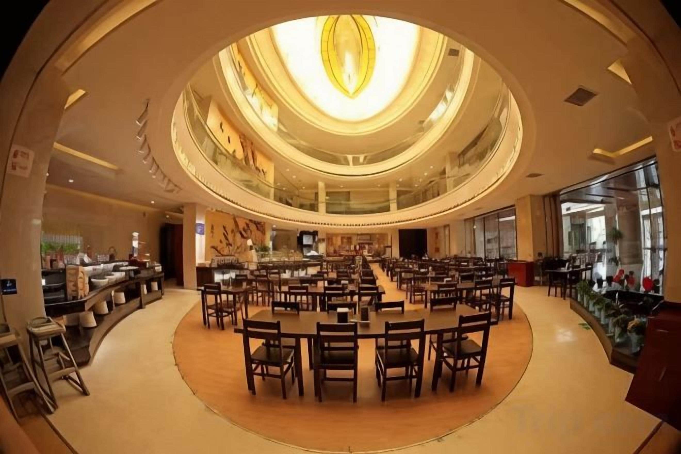 A spacious indoor area with multiple dining tables and chairs arranged in a circular pattern, a central light fixture, and a glass ceiling.