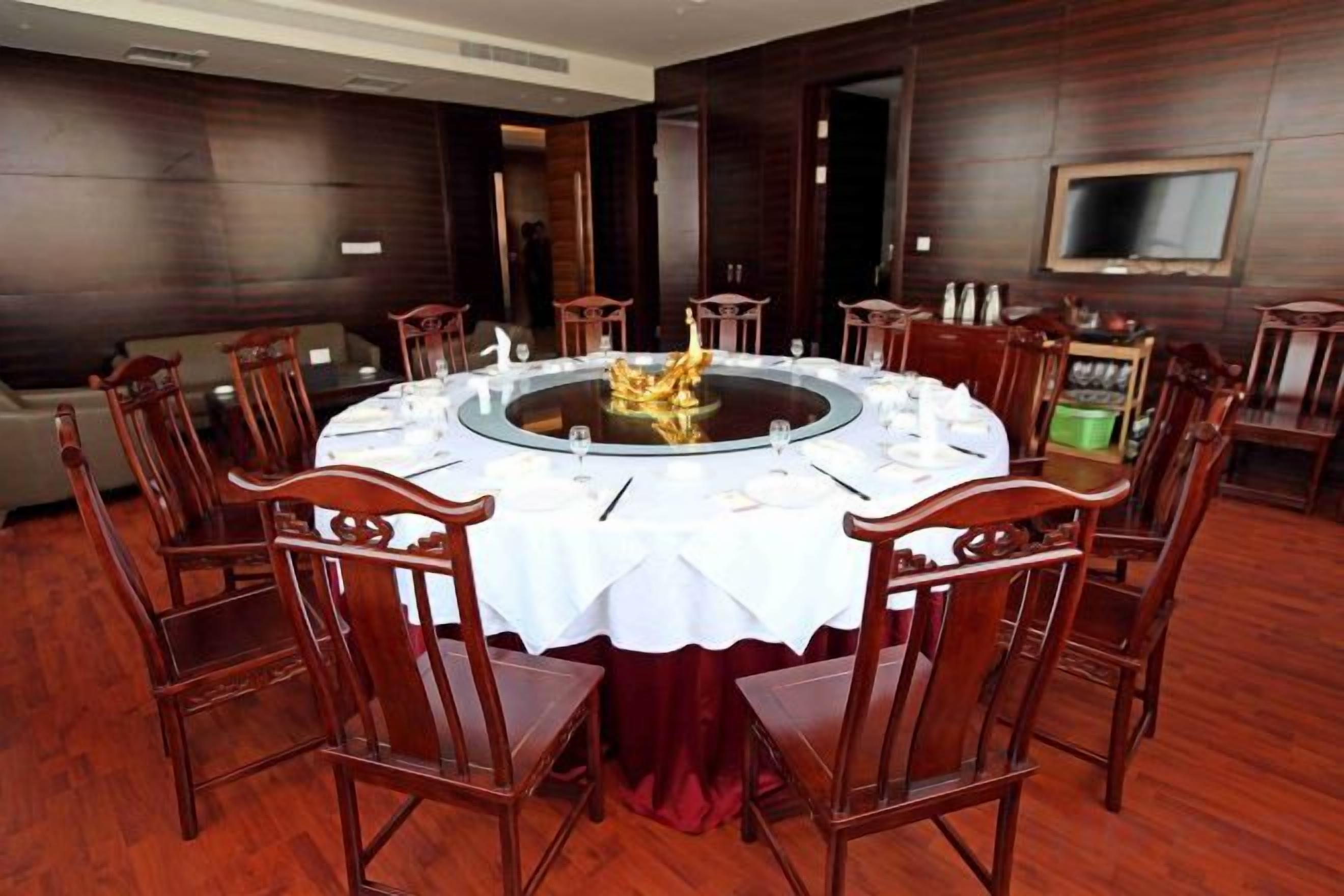 A round dining table with a white tablecloth, surrounded by wooden chairs, in a room with dark wooden walls and a flat-screen TV mounted on the wall.