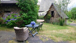 A stone cottage with a thatched roof, a wicker chair, a table with a vase of flowers, and a small garden with purple flowers.