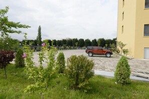 Un área de estacionamiento con un vehículo rojo, rodeada de vegetación y un edificio de fachada amarilla.