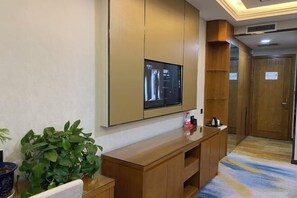 Een hotelkamer met een grote flatscreen televisie aan de muur, een houten console tafel, een potplant en een deur met een bordje.