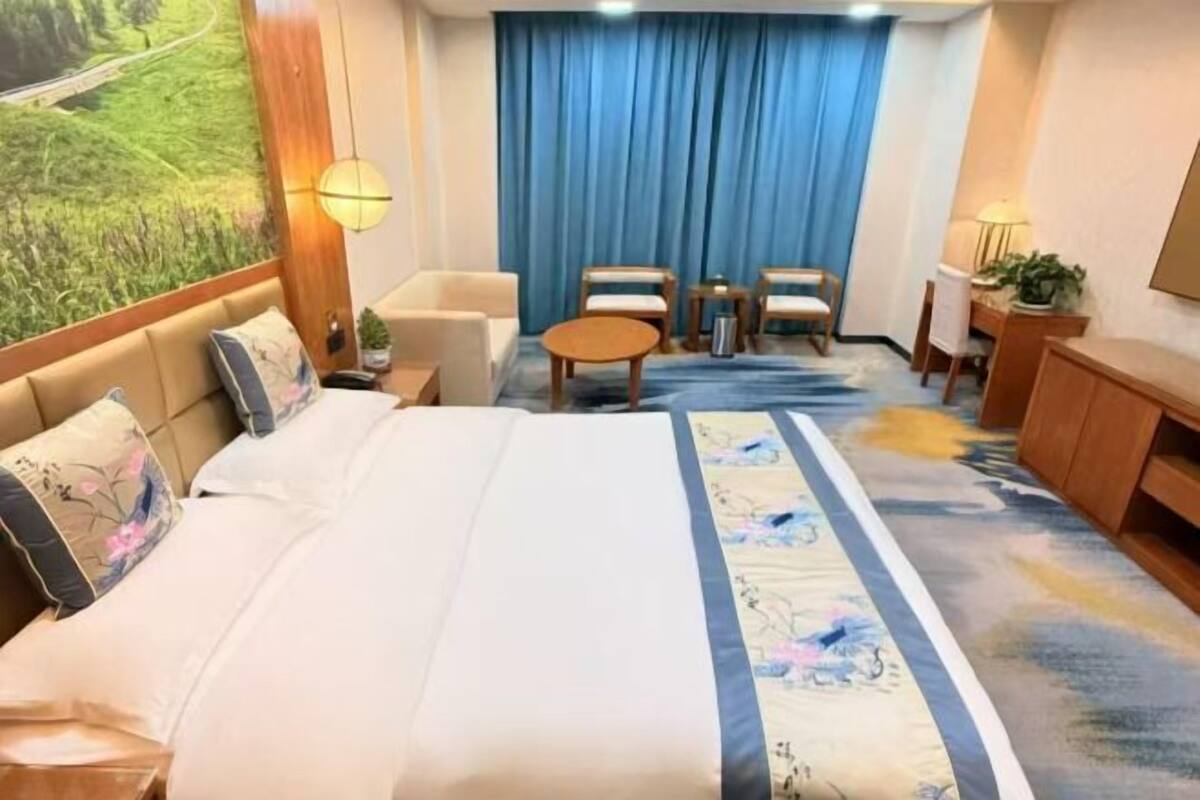 Una habitación de hotel con una cama grande, una mesita redonda pequeña, dos sillas, un escritorio y un ventanal con cortinas.