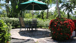An outdoor patio area with stone flooring, a table and chairs, a green umbrella, and lush greenery including a flowering bush.