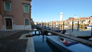 A canal with boats docked, buildings with windows, and a tower in the background. - Biennial-Pulse (Venezia)