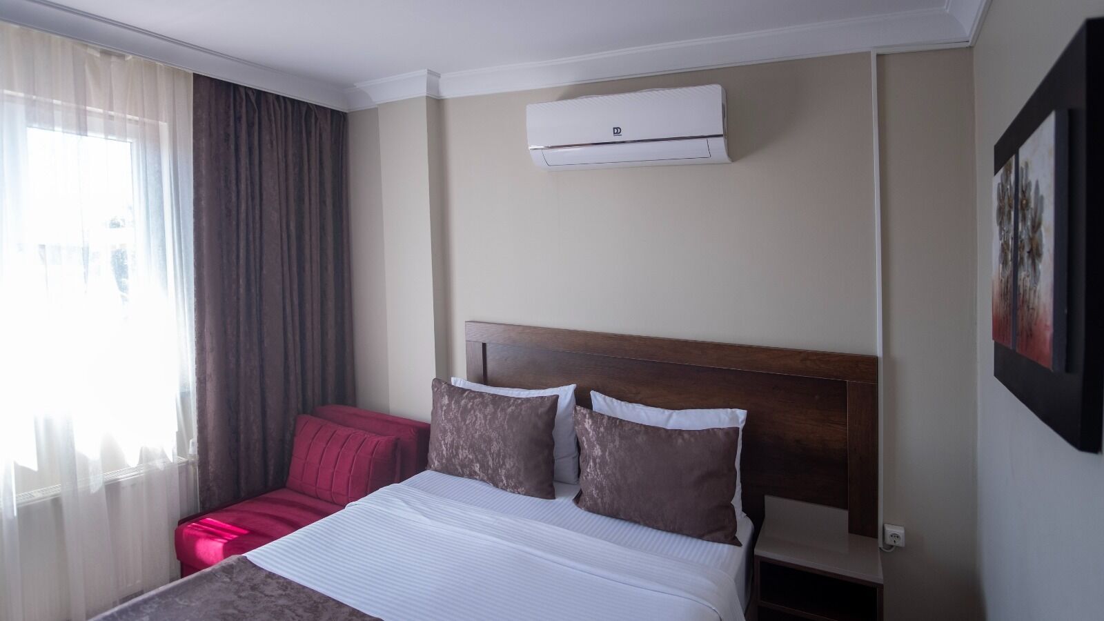 A hotel room with a bed, a red chair, a window with curtains, and an air conditioning unit.