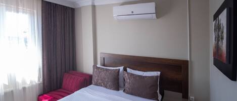 A hotel room with a bed, a red chair, a window with curtains, and an air conditioning unit.