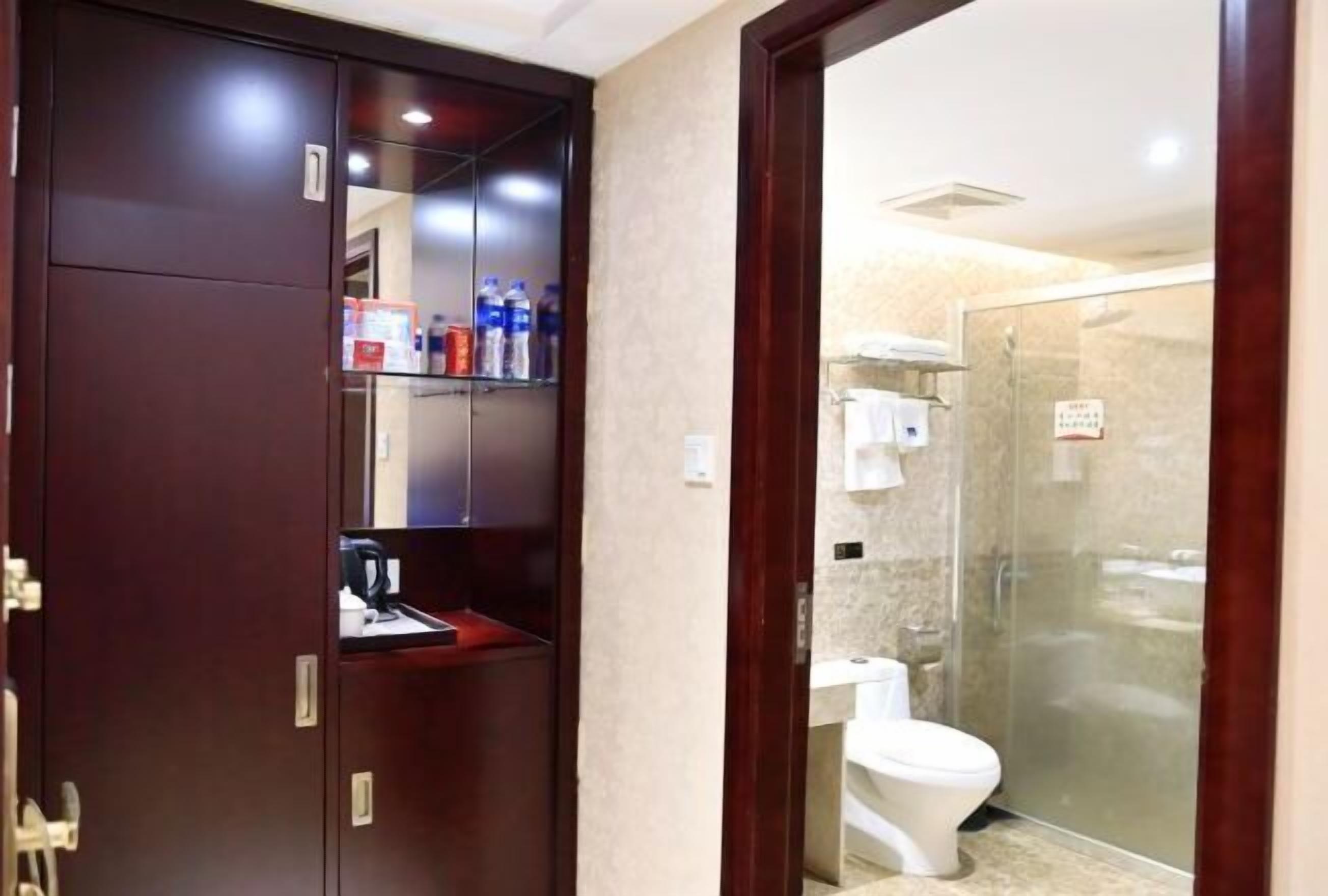 A hotel bathroom with a glass shower enclosure, a white toilet, and a dark wooden cabinet with shelves.