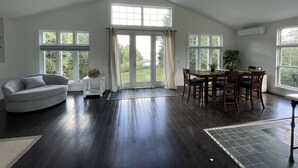 A spacious room with a high ceiling, a dining area with wooden chairs and a table, a gray armchair, and large windows offering a view of the outdoors.
