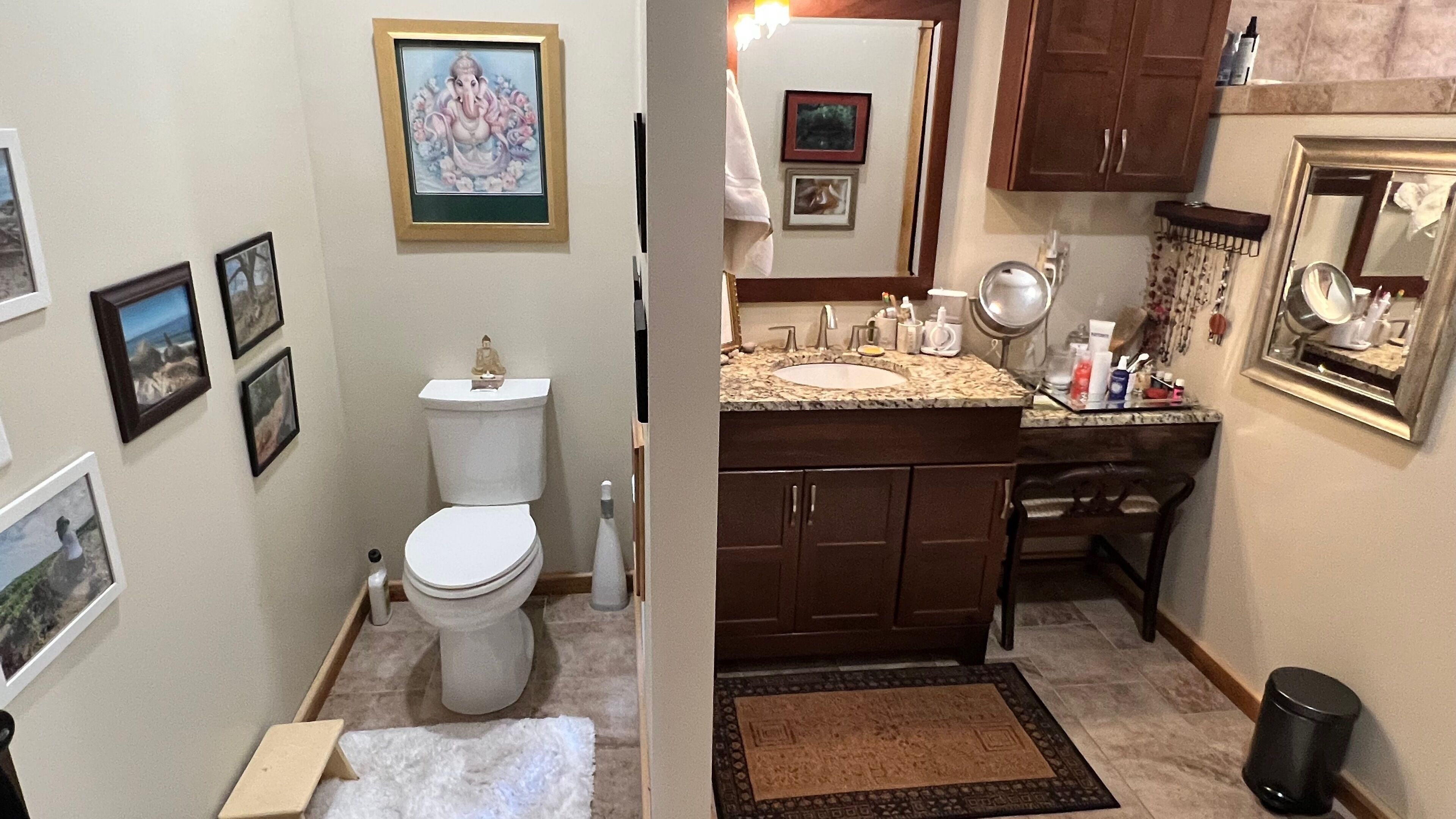 A bathroom with a toilet, sink, and mirror. There are framed pictures on the wall and a small Buddha statue on the toilet tank.