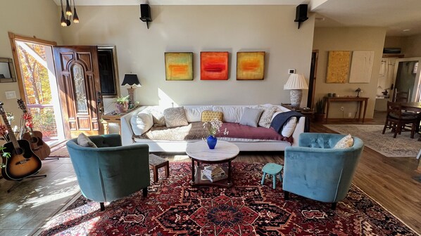 Ein Wohnzimmer mit einer Couch, zwei Sesseln, einem Couchtisch und einem gemusterten Teppich. An der Wand hängen drei Bilder, eine Gitarre steht dort, und ein Fenster bietet Blick auf Bäume.