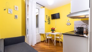 Et kompakt værelse med en sofa, et lille spisebord med stole, et tekøkken med komfur og et tv ophængt på væggen.
