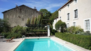 A swimming pool with a wooden deck and lounge chairs, surrounded by greenery and a large building in the background.