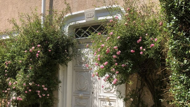 A white door with a decorative design, surrounded by lush greenery and pink flowers.
