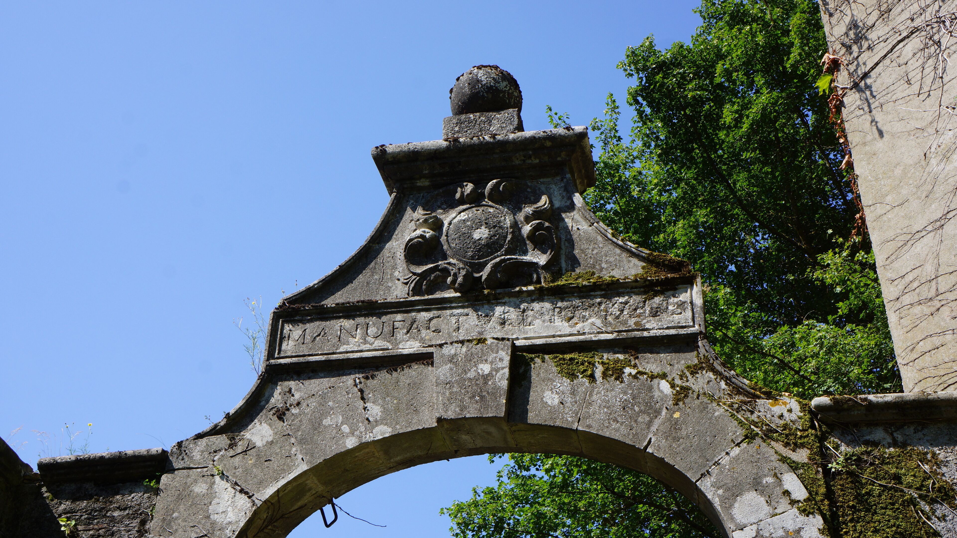 An old stone archway with a decorative crest and inscriptions, surrounded by greenery.