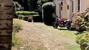 A stone archway leading to a garden with a motorcycle parked near a building with a window and a door.