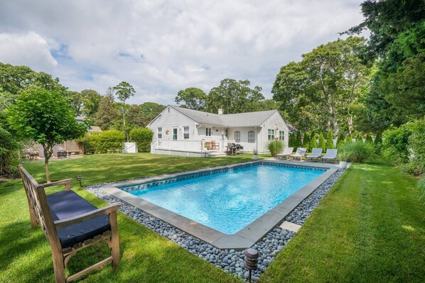 A backyard with a swimming pool, a wooden deck, and a white house. - Chic modern beach cottage with heated pool (Hampton Bays)