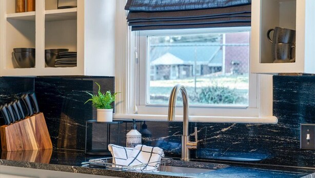 Una cocina con encimera de mármol negro, pileta de acero inoxidable y ventana con vista a una casa y follaje.