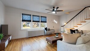 Amplia sala de estar con piso de madera, un sofá gris, una mesa de centro, un mueble para televisión y una escalera con barandilla negra.