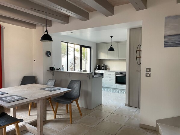 A modern kitchen with white cabinets, a dining area with a wooden table and chairs, and a view of the outside through a window. - Charming Ile de Ré house, 2 steps from beaches and golf course. (Saint-Clément-des-Baleines)