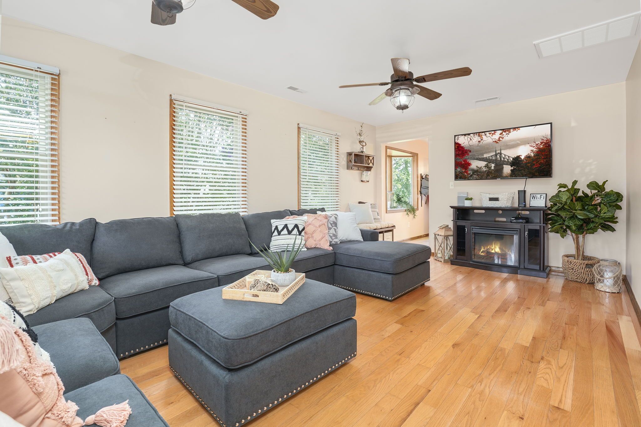 A living room with a sectional sofa, ottoman, wooden flooring, ceiling fan, fireplace, TV, and large windows with blinds.