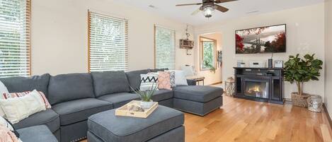 A living room with a sectional sofa, ottoman, wooden flooring, ceiling fan, fireplace, TV, and large windows with blinds.