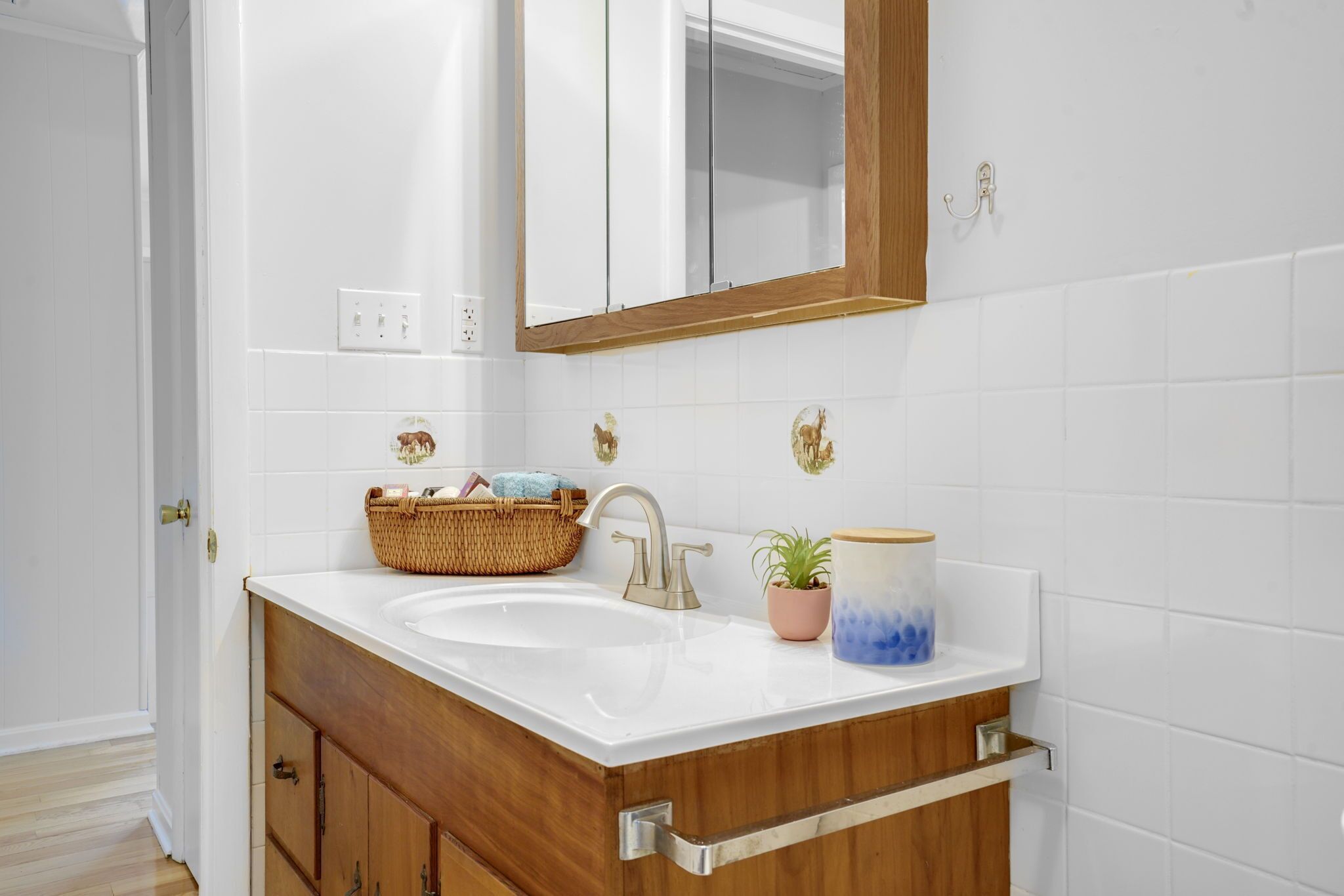 A bathroom with a white sink, wooden vanity, and a mirror. There is a basket on the sink, a potted plant, and a decorative container.